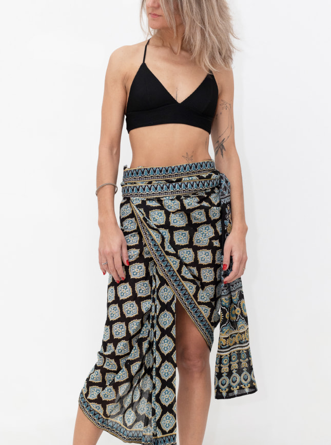 The black and blue blockprint wrap dress is worn as a wrap around skirt. Worn on the hips it is tied around the waist to give an asymetric skirt. The model is wearing her own black crop top with the skirt. Perfect to wear with a bikini it is a deluxe sarong.