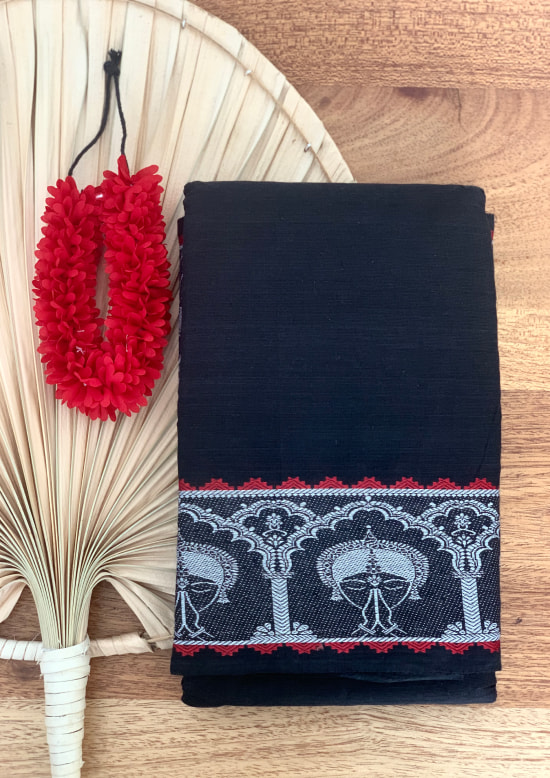 The ByGungur Anjali Saree is a black saree with a white and red border. The border is embroidered in white with the face of an Odissi dancer holding her hands in Anjali mudra. The dancer's face is surrounded by a temple inspired motif. The saree is made of 100% cotton and can be worn as an Indian classical dance practice saree or for a formal occasion.