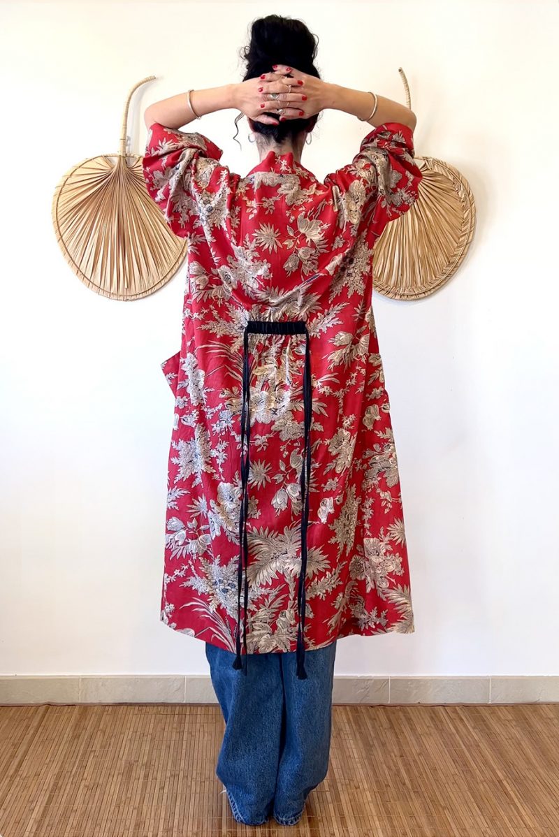 The Lily Kimo is a 100% cotton kimono jacket. It has pockets and a thin, black, adjustable belt attached at the back of the garment. Here we see the back view. The model is wearing the floral kimono with jeans.