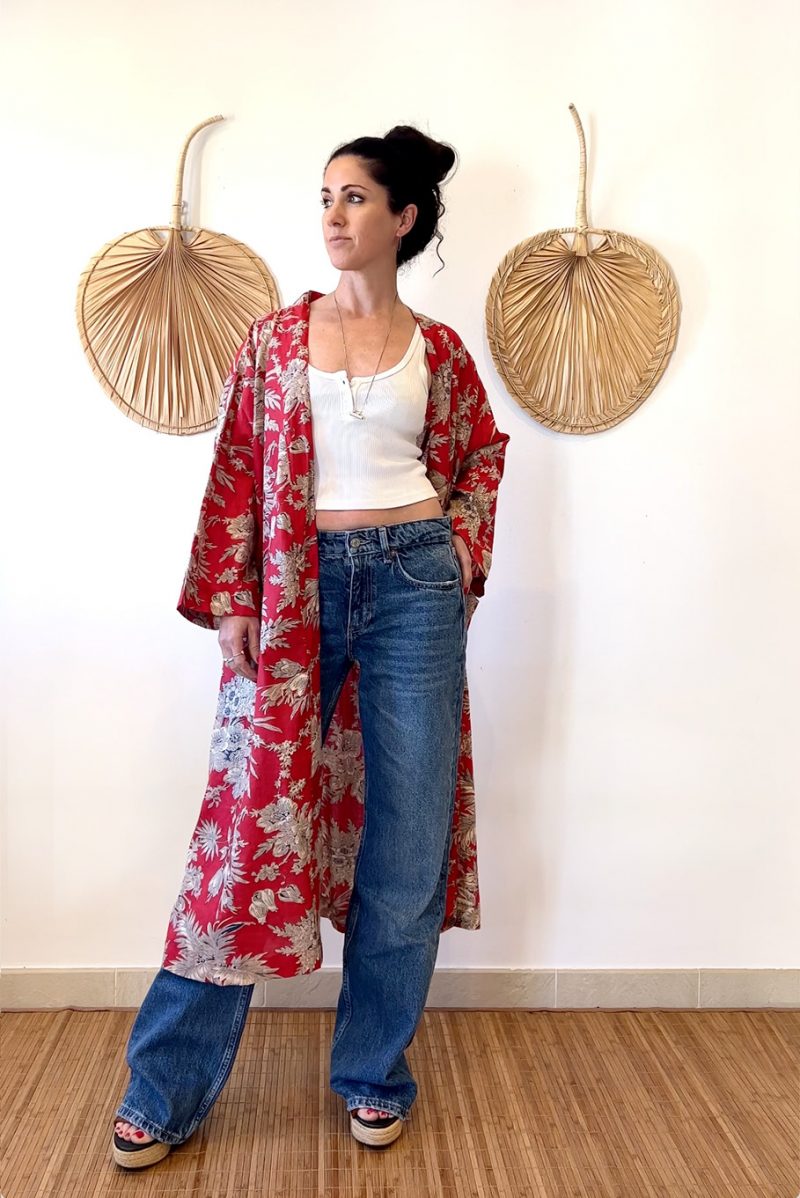 The Lily Kimo is a floral print cotton kimono jacket with pockets and a thin, black, adjsutable belt attached at the back. It is a cream floral design on a red backgroundHere the model is wearing the kimono with jeans and a white top. The Lily Kimo can be worn casual or smart for a distinctive look.